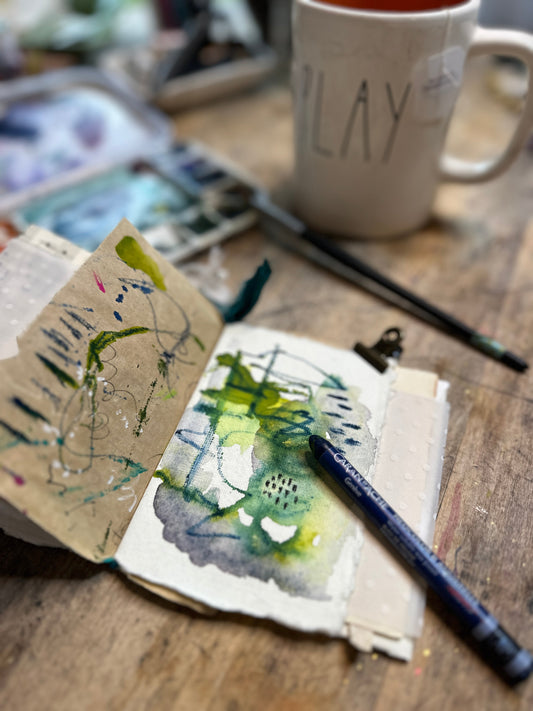 Mixed-media art journal with colorful marks and art supplies on a wooden studio table.